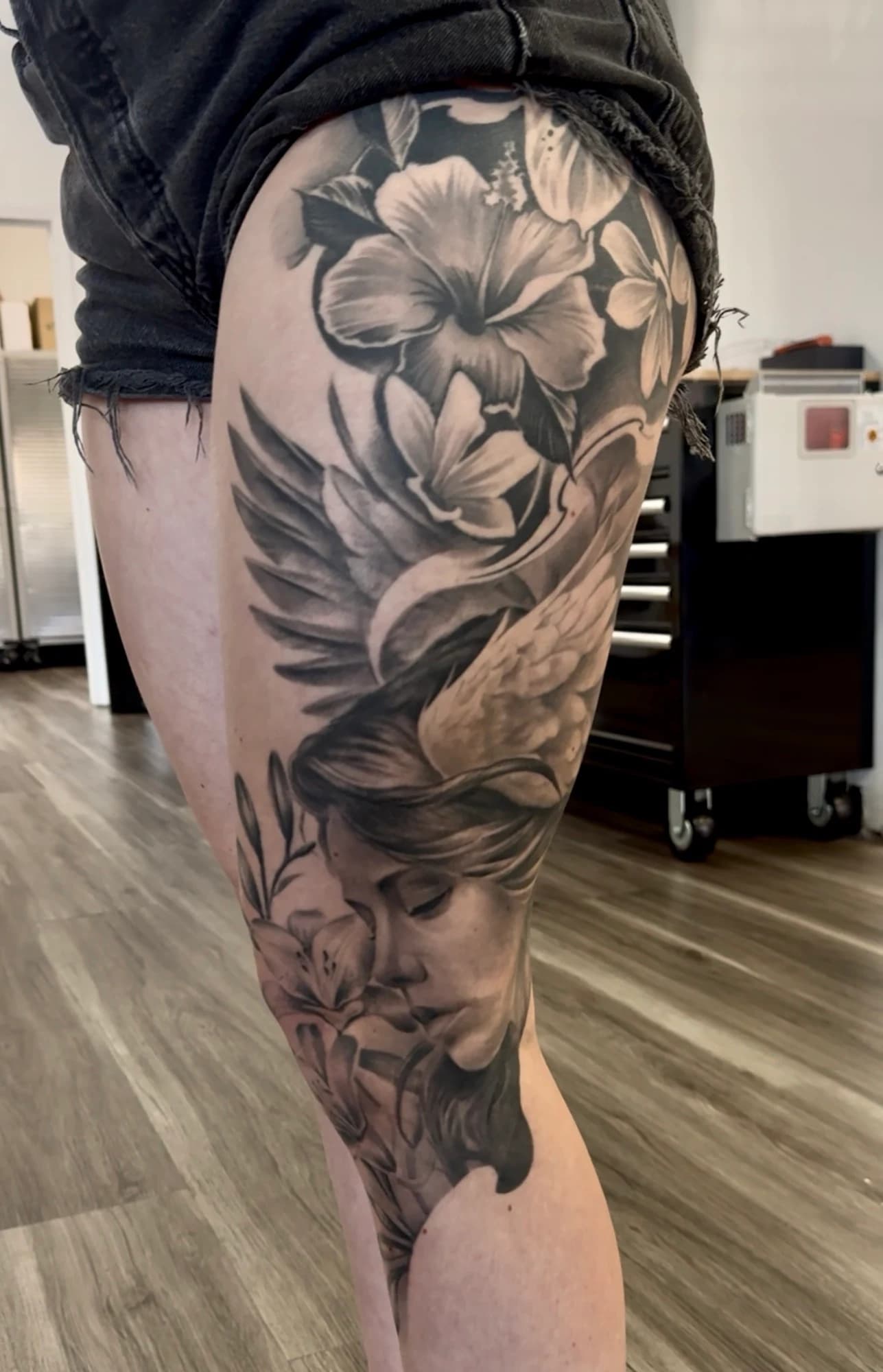 Black and grey tattoo: flowers, a bird, and a portrait flowing down the leg.