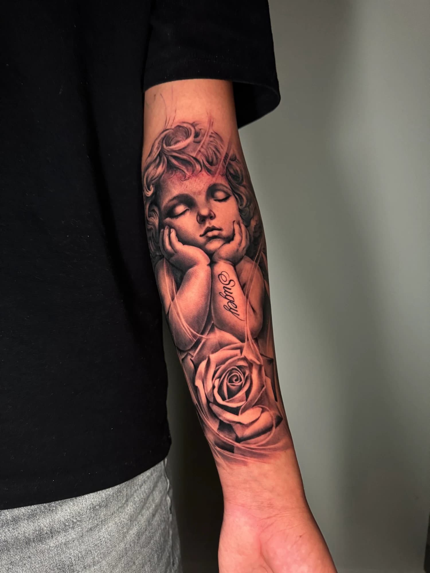 Black and grey tattoo: cherub and rose with soft shading and fine detail.